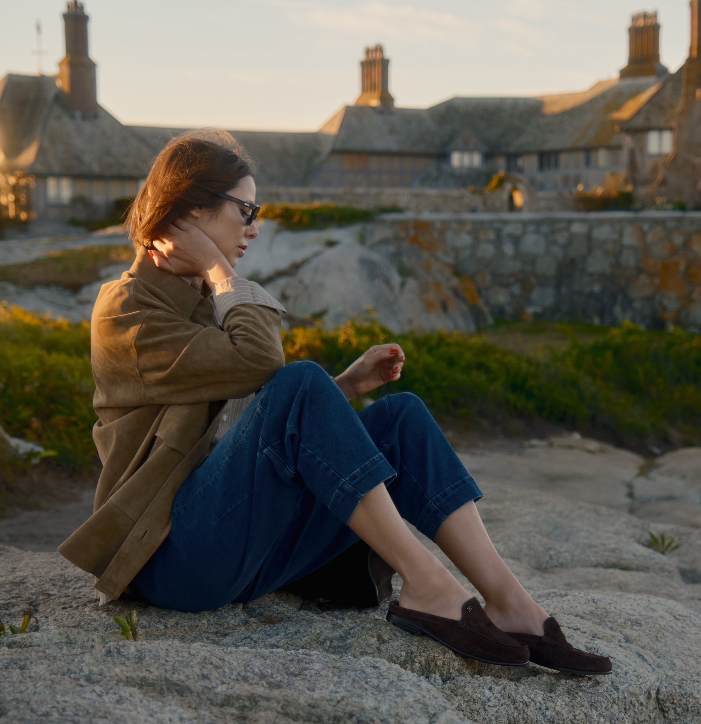 Woman sitting outdoors on a rock with stone buildings in the background. She is wearing a tan suede jacket, cuffed blue jeans, and the Franco Sarto Mavis Mule Loafer in dark brown suede.