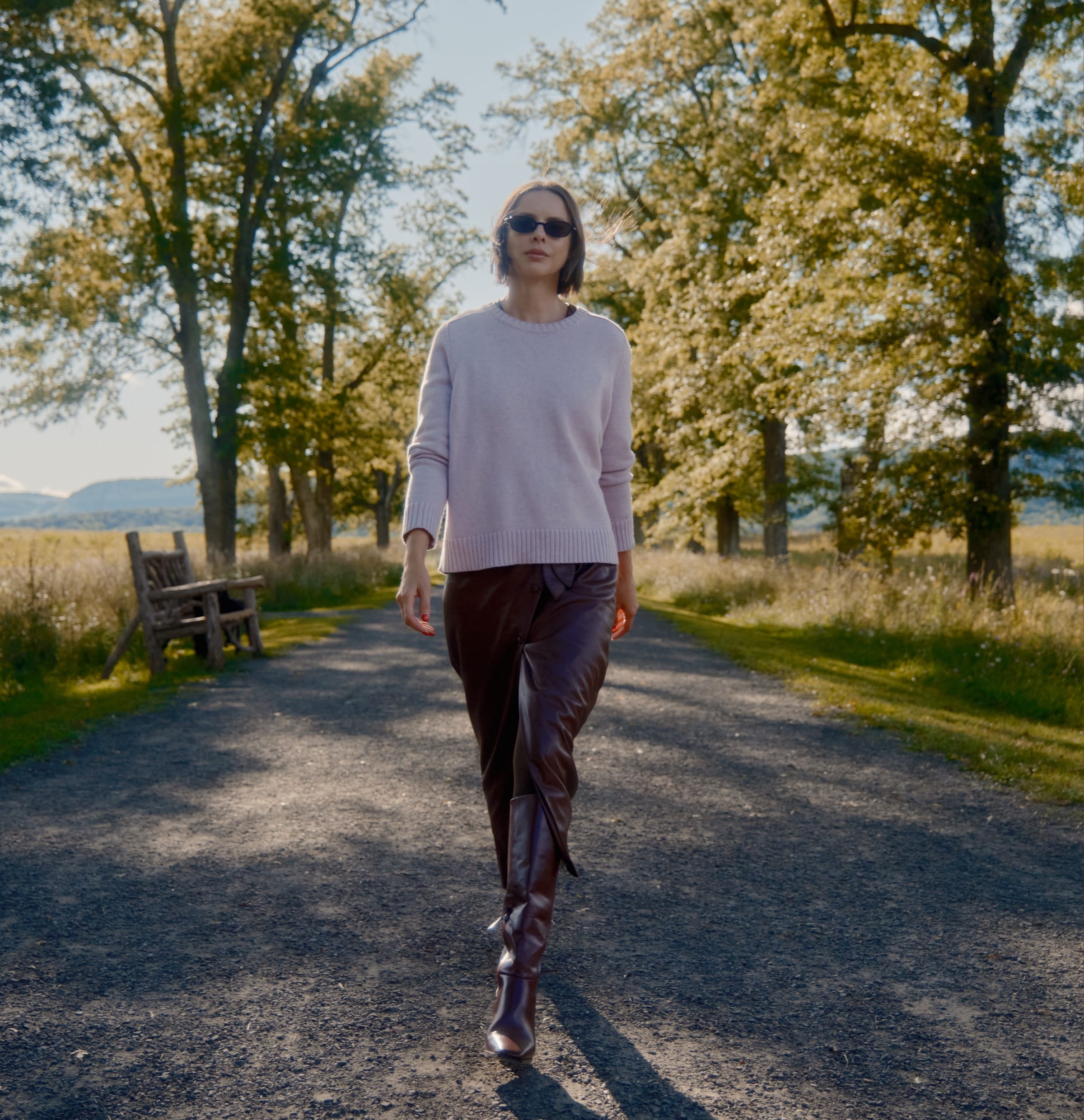 Woman walking down a tree-lined path wearing a light pink sweater, dark brown leather pants, and Franco Sarto’s Jonie Knee High Boot in oxblood red leather.  