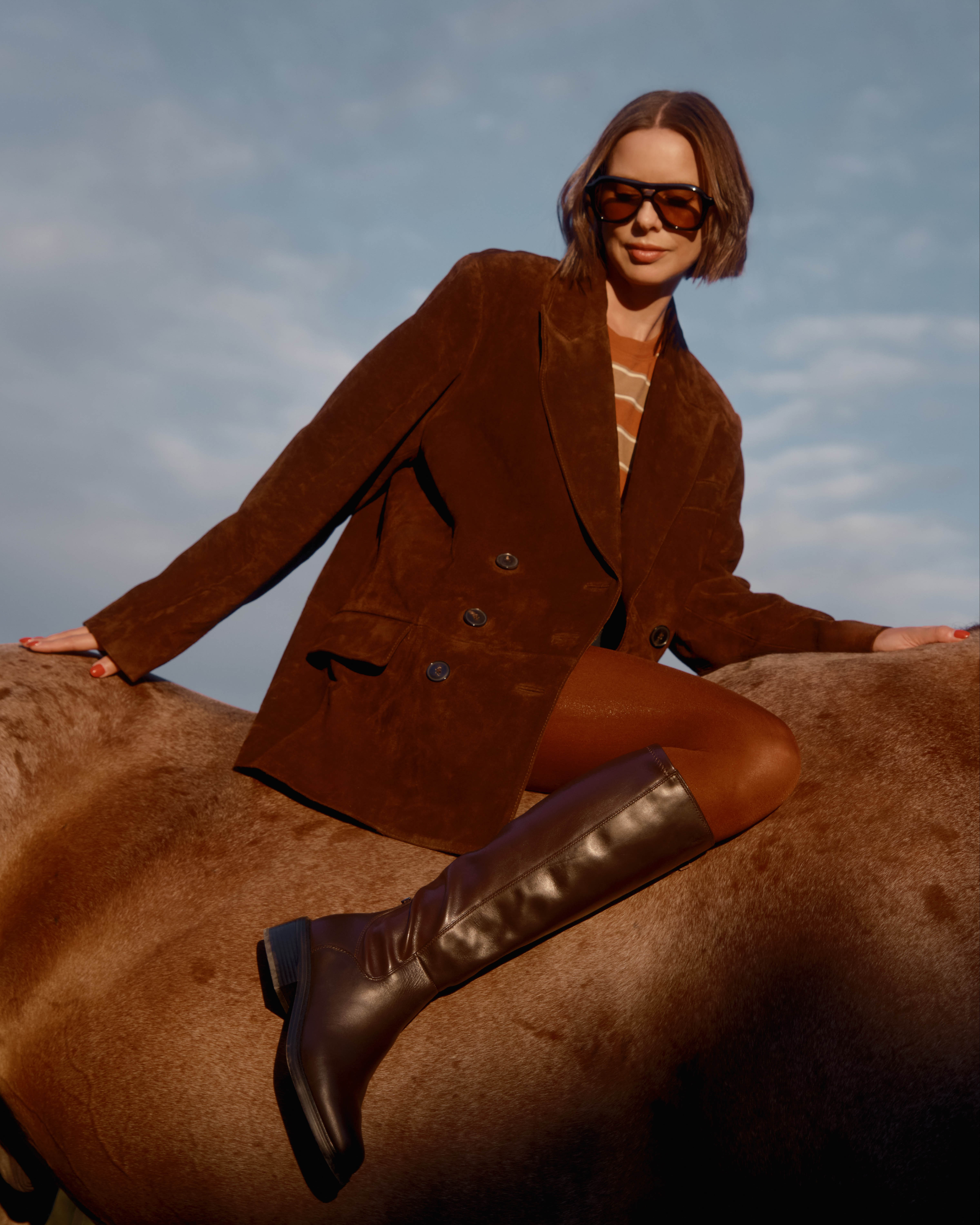 Woman sitting on a horse wearing a brown blazer, sheer brown tights, and the Franco Sarto Gisella Knee High Boot in brown leather.  