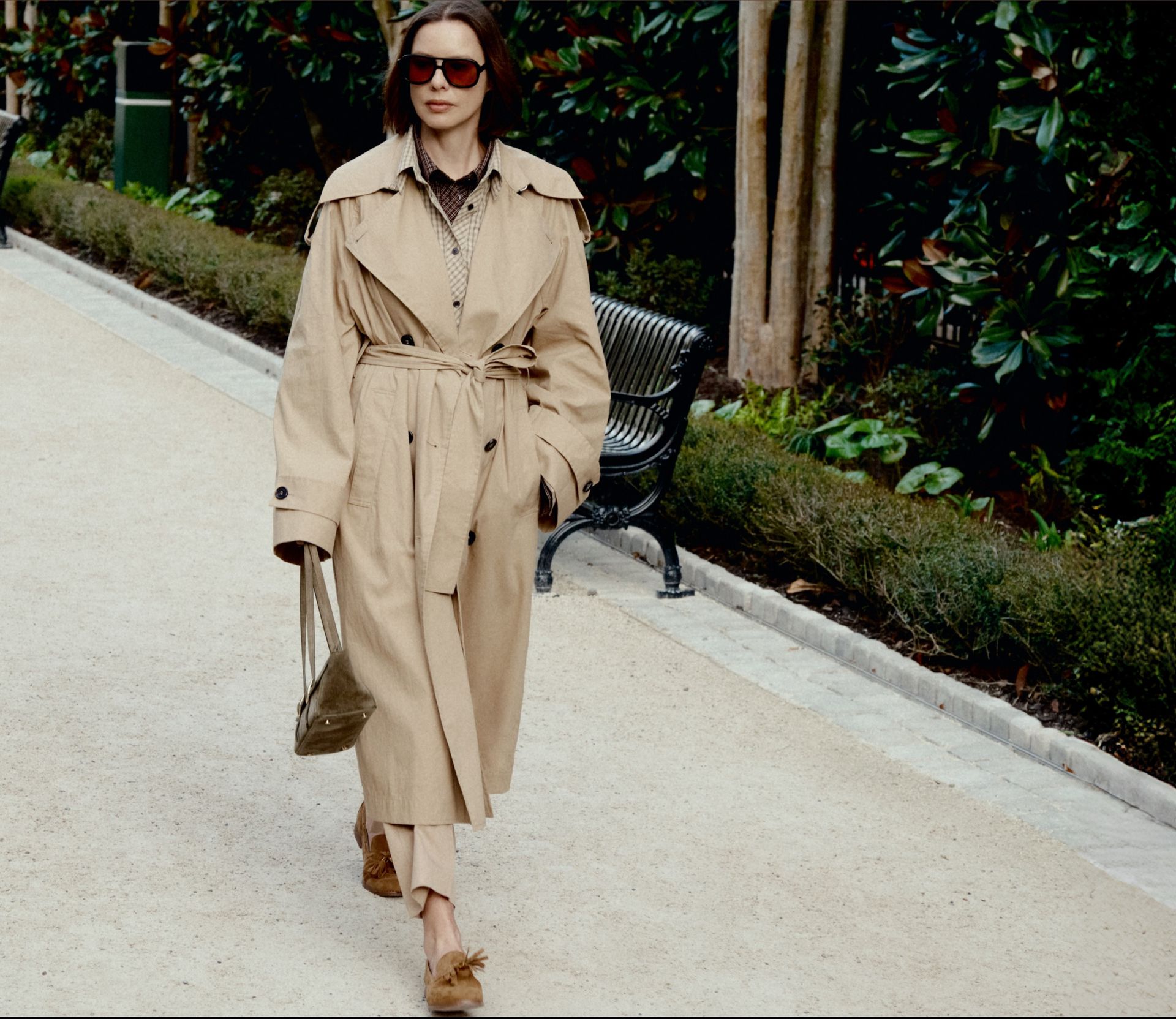 Woman walking in a garden path wearing Franco Sarto Hartlie Tassel Loafer in tan suede with a belted trench coat.