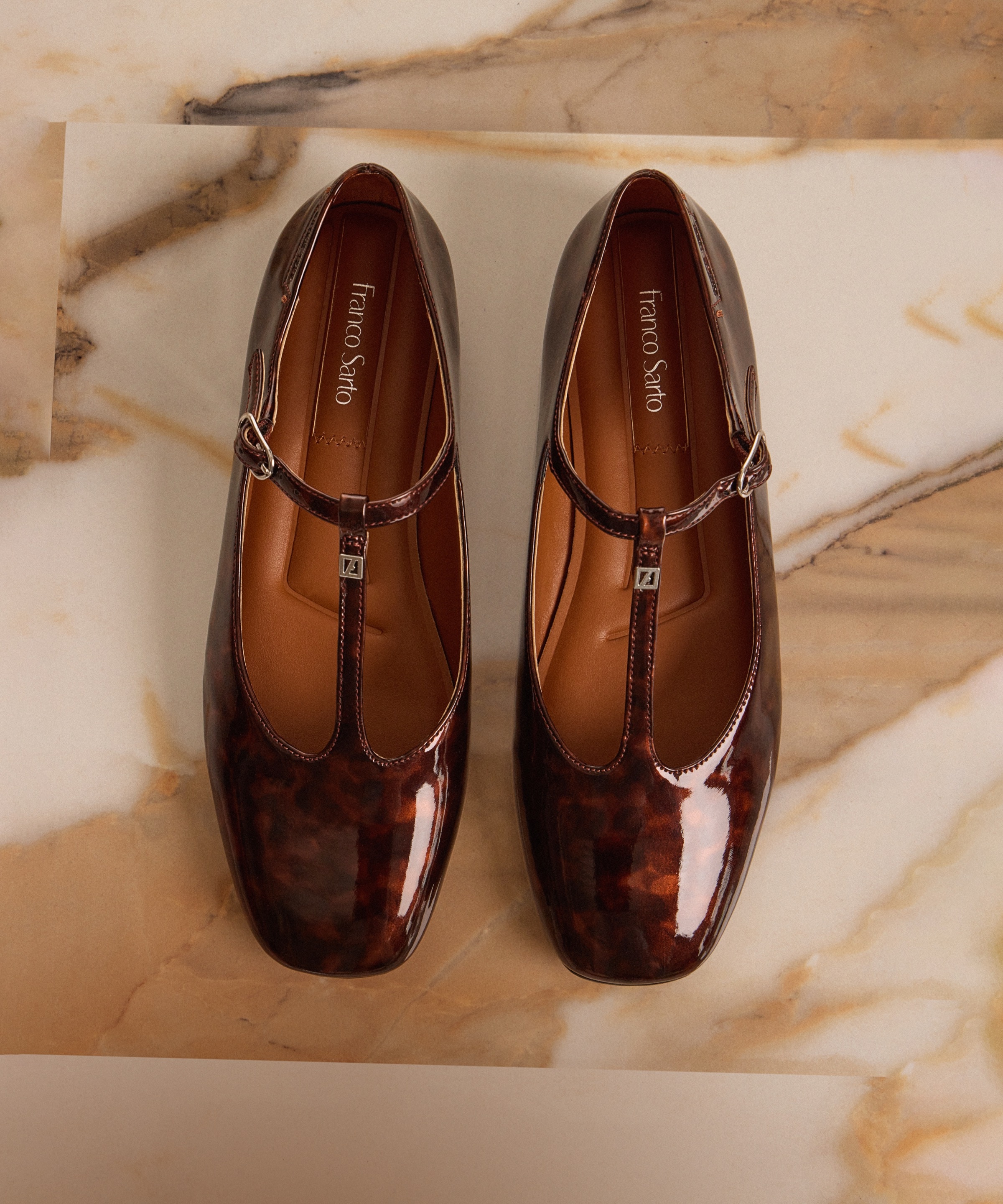 Top view of a pair of women’s Marylee Mary jane flats in tortoise-print patent leather displayed on a marble background.  