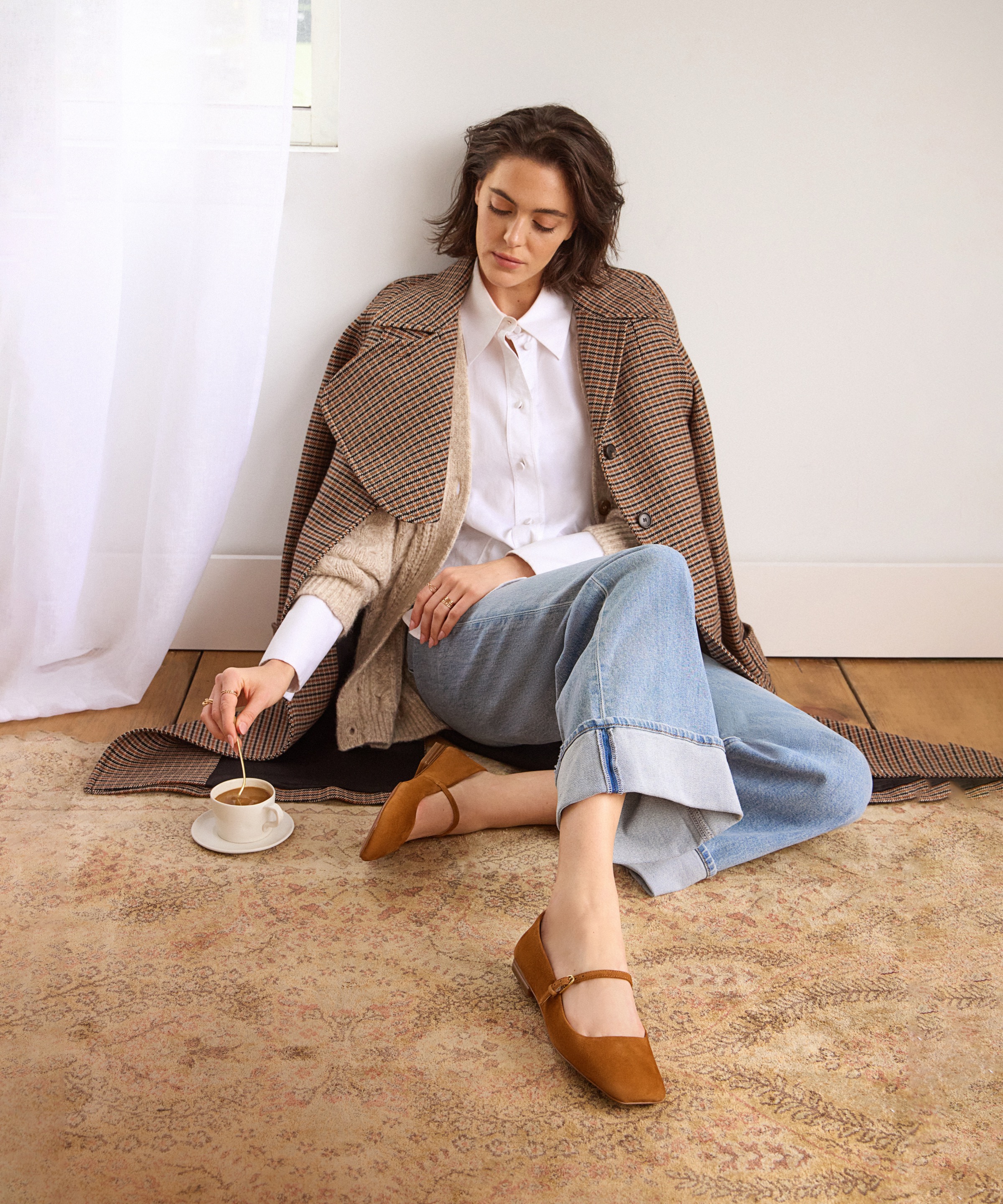 A woman wearing a pair of brown Tinsley Mary Jane flats sitting on the floor stirring a cup of coffee.   