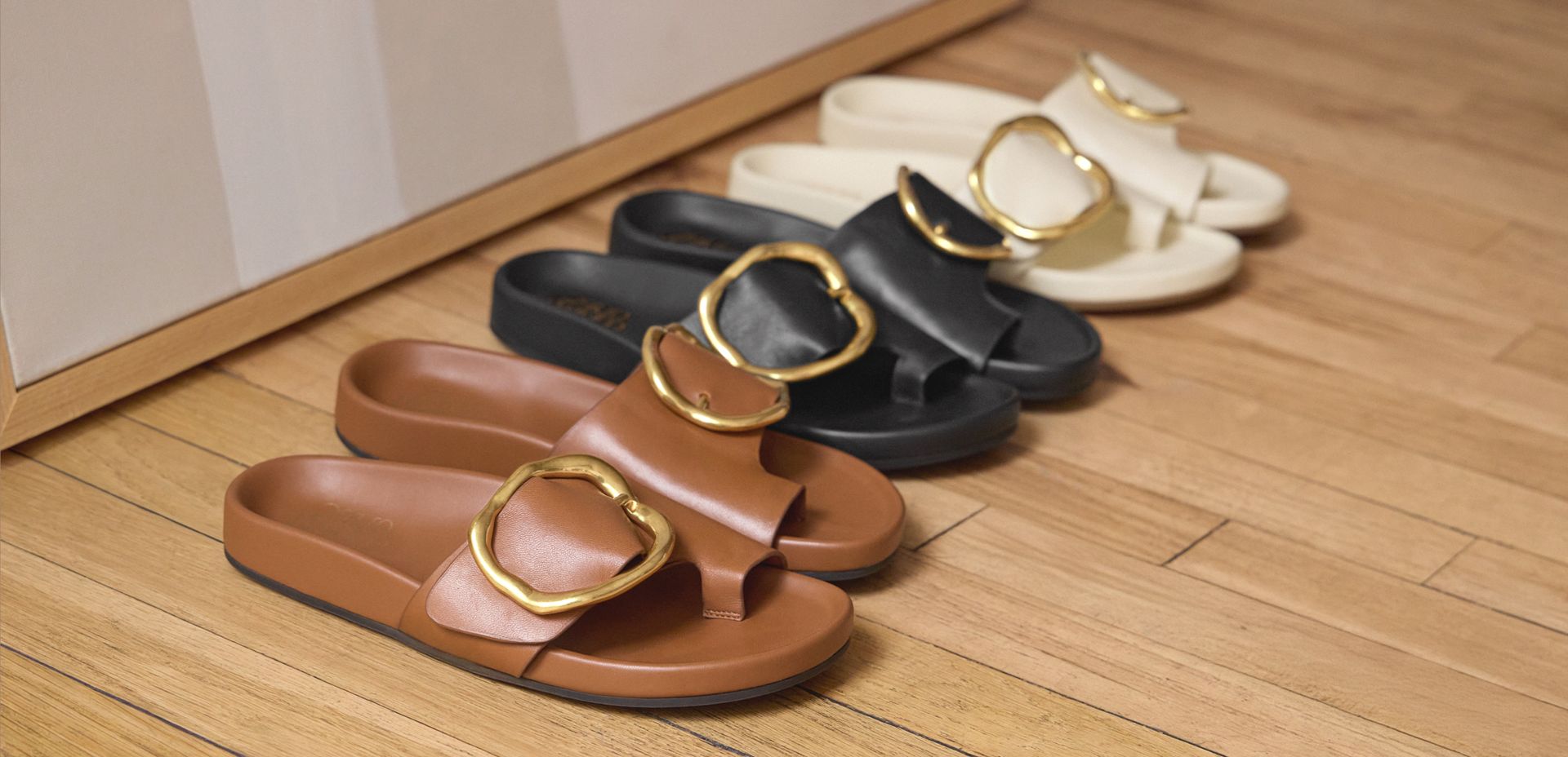 Franco Sarto Brianka Slide Sandals in brown, black, and ivory leather with gold buckle detail, lined up on hardwood flooring