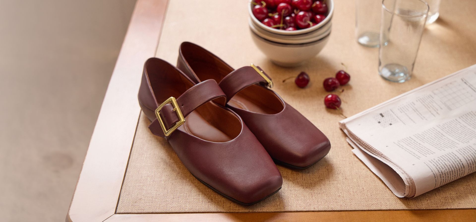 Franco Oakes Mary Jane in Bordo red leather, single strap with gold buckle, styled on a table with a bowl of cherries and a newspaper.