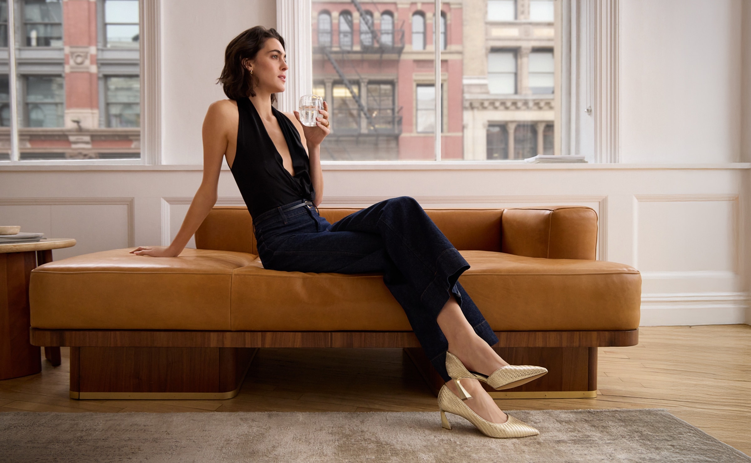 a woman relaxing on the couch wearing a chic outfit and the the tawny pump in gold faux leather