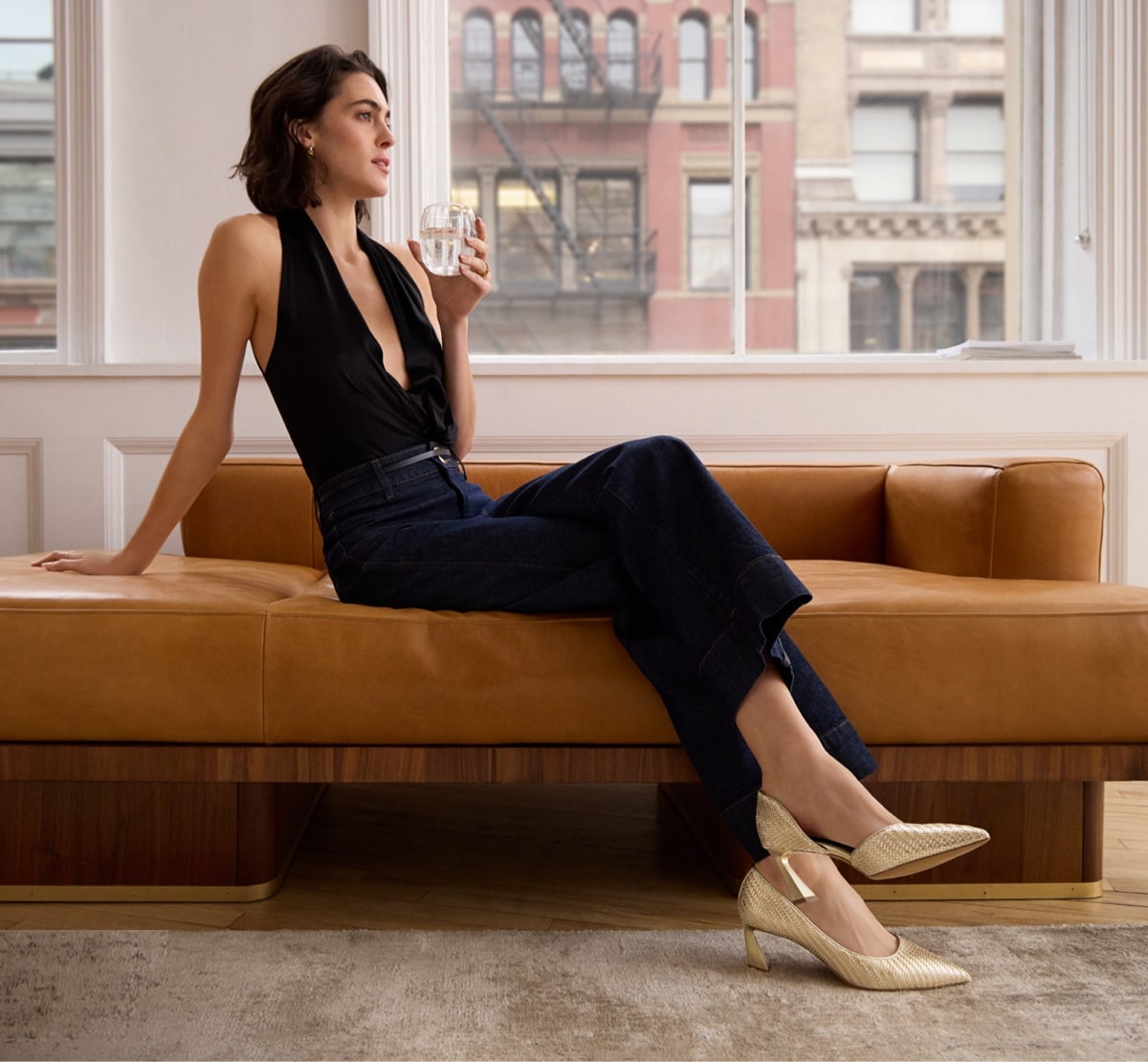 a woman relaxing on the couch wearing a chic outfit and the the tawny pump in gold faux leather